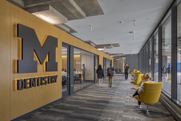 A wide hallway with people sitting in bright yellow chairs next to the window